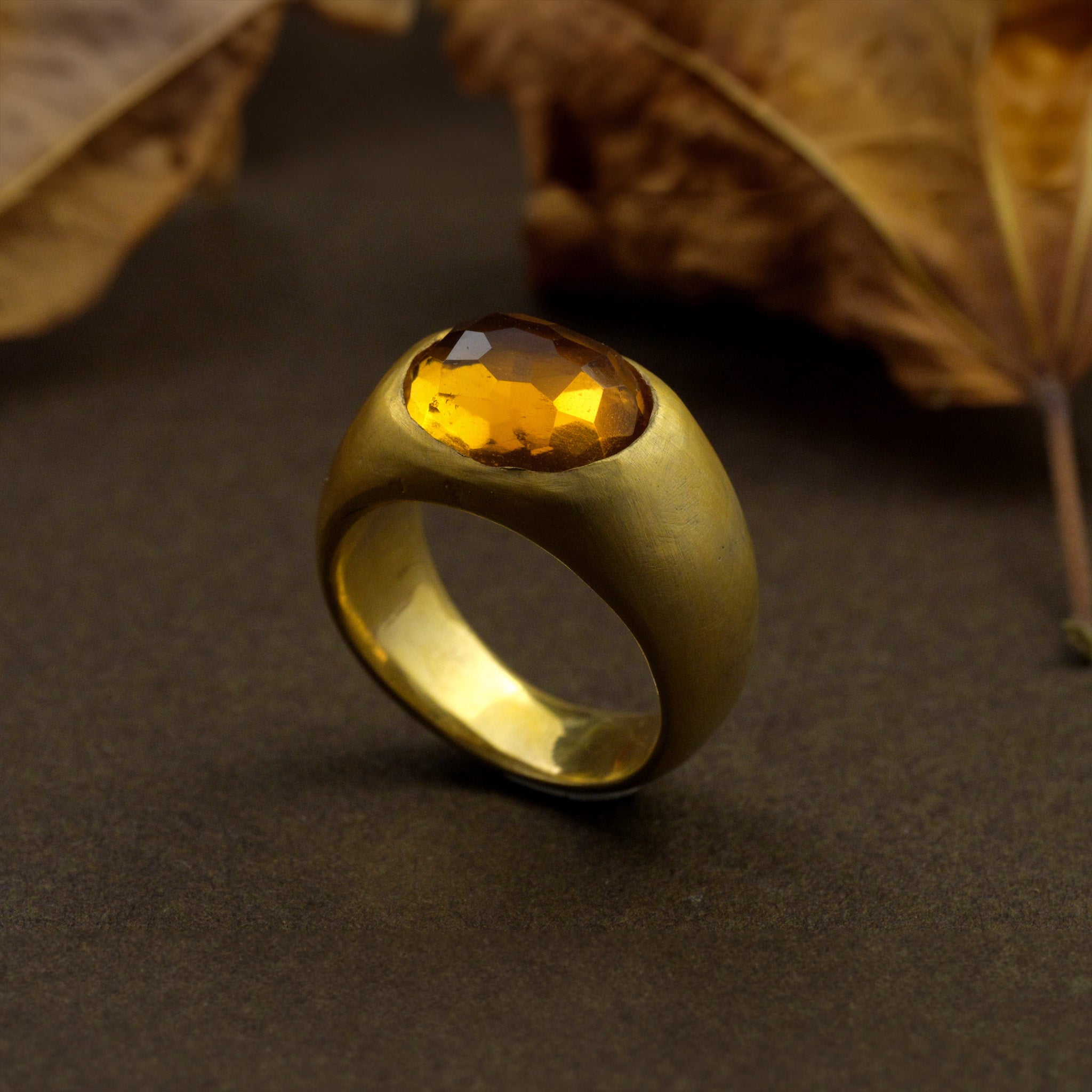 A timeless gold oval ring adorned with a captivating Citrine gemstone on a dark surface with dry leaves in the background. This classic piece exudes traditional elegance and is beloved for its enduring appeal.