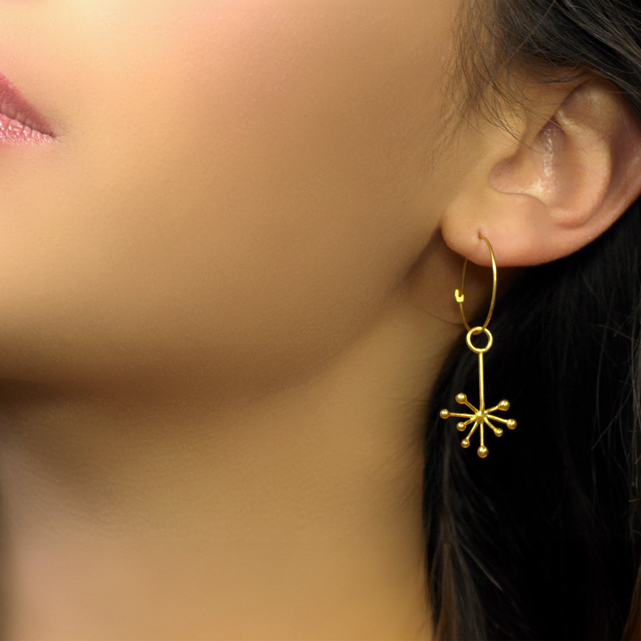 Close up of delicate 18k yellow gold earrings on a model, featuring an intricate snowflake design formed by thin, radiating gold lines. The design exudes a subtle yet playful aesthetic.