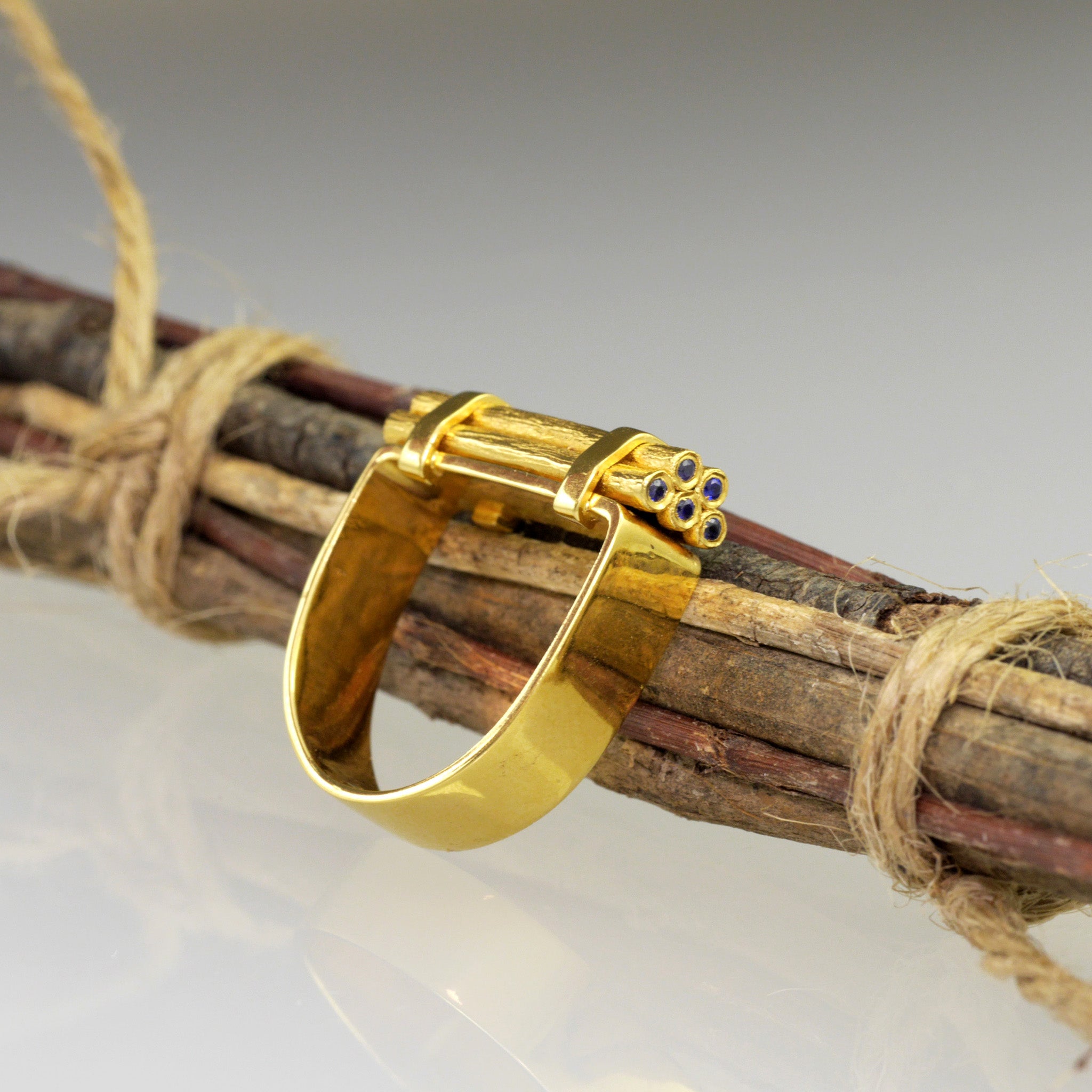 Handcrafted gold ring with stacked wood-inspired design, adorned with sparkling Sapphires for added color and radiance. the ring lies on a pile of stacked wood.