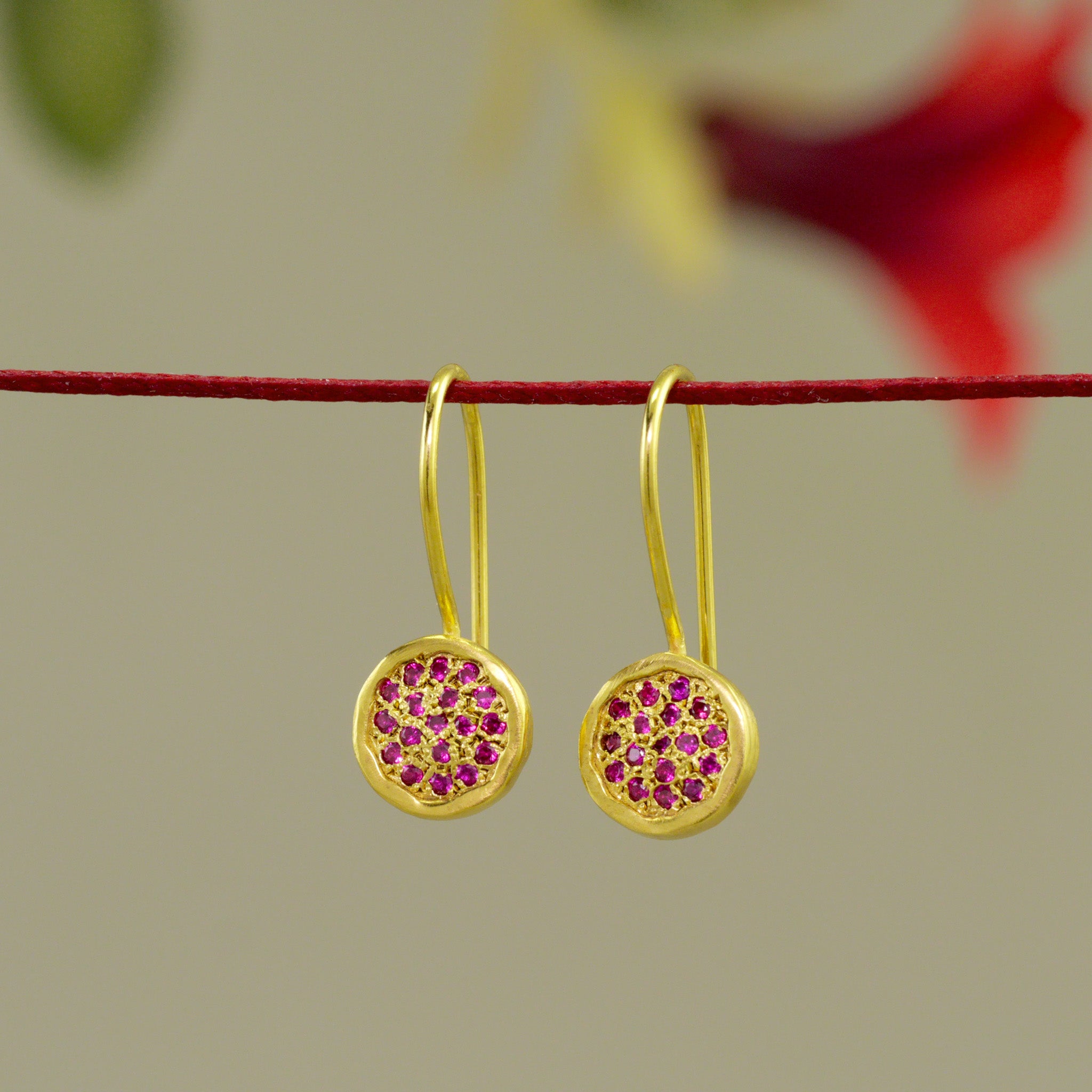 Close up of 18k Gold drop earrings adorned with twinkling ruby gemstones hanging from a red thread and red flower in the background.