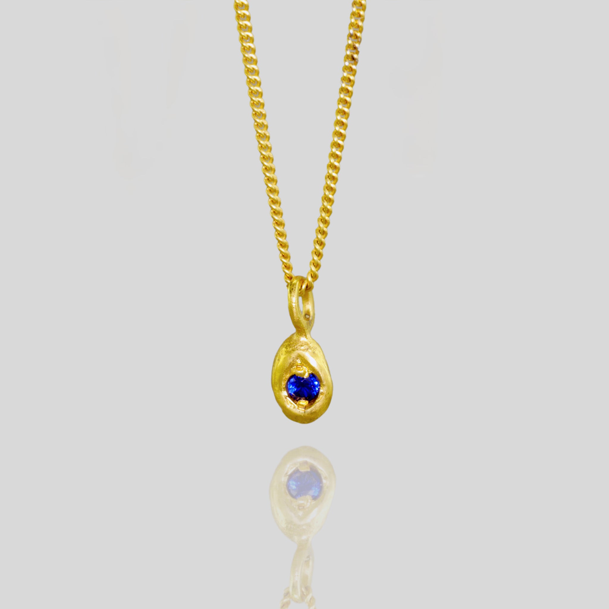 close up of an 18k Yellow Gold Pendant with a drop shape and a central sapphire, designed to replicate a naturally gathered dried seed.