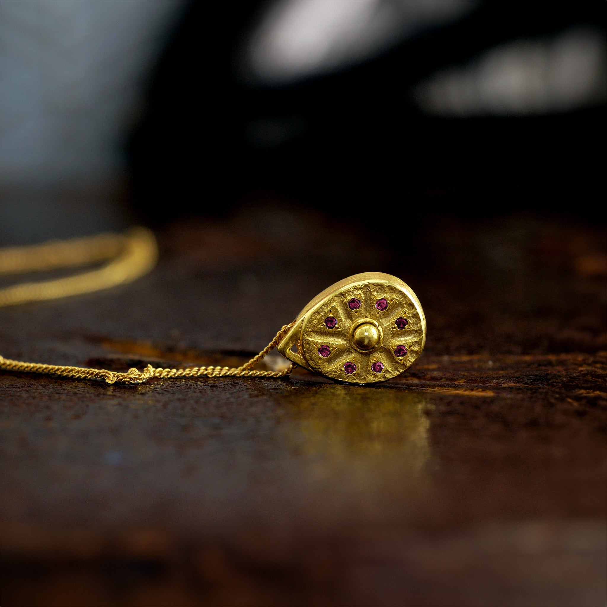Close up of 18k Gold pendant resembling a Sundial clock, accented with small Rubies between the edges of each clock hand on a dark rusty background. A luxurious and timeless addition to any collection.
