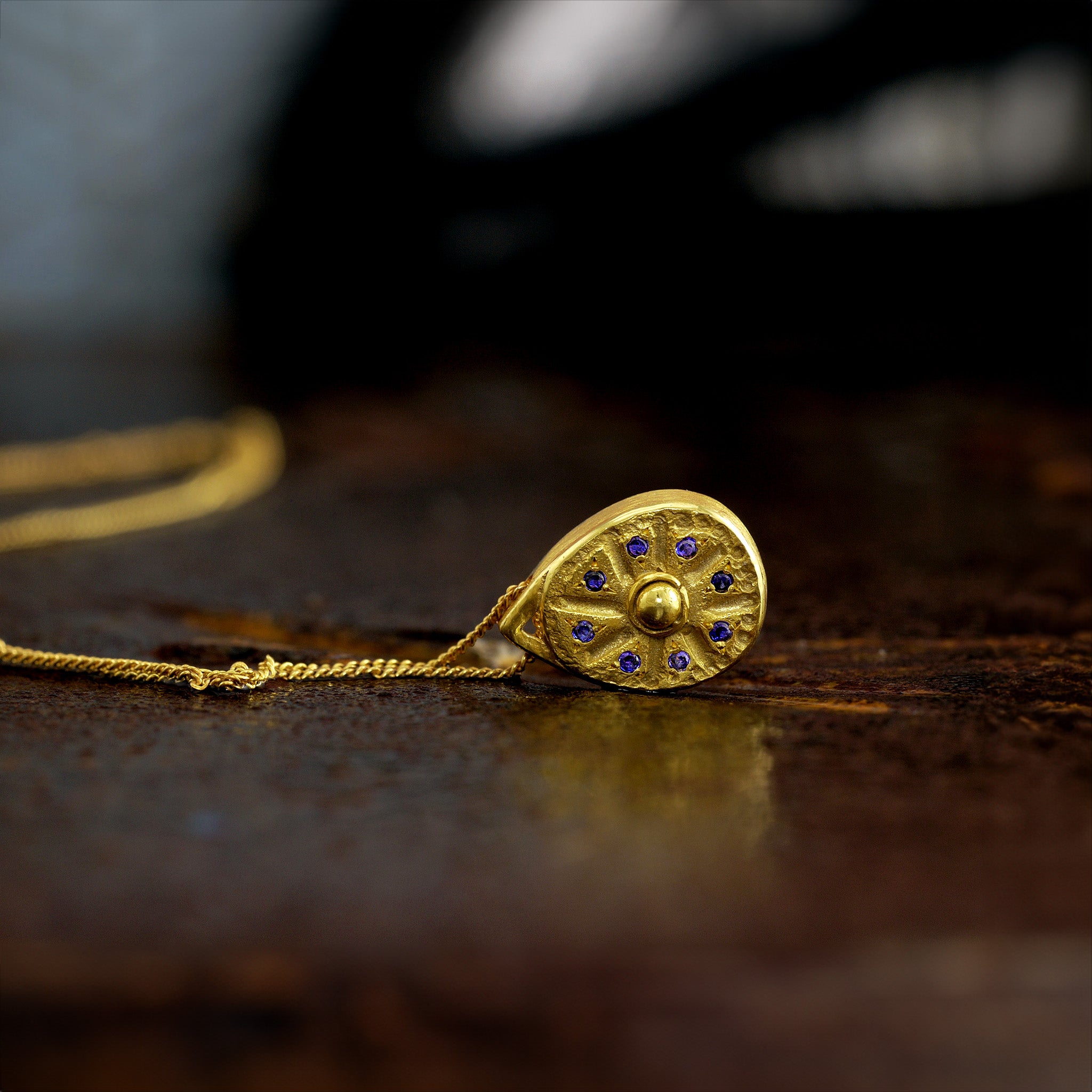 Close up of 18k Gold pendant on a dark rusty background. The pendant resembles a Sundial clock, accented with small Sapphires between the edges of each clock hand. A luxurious and timeless addition to any collection.