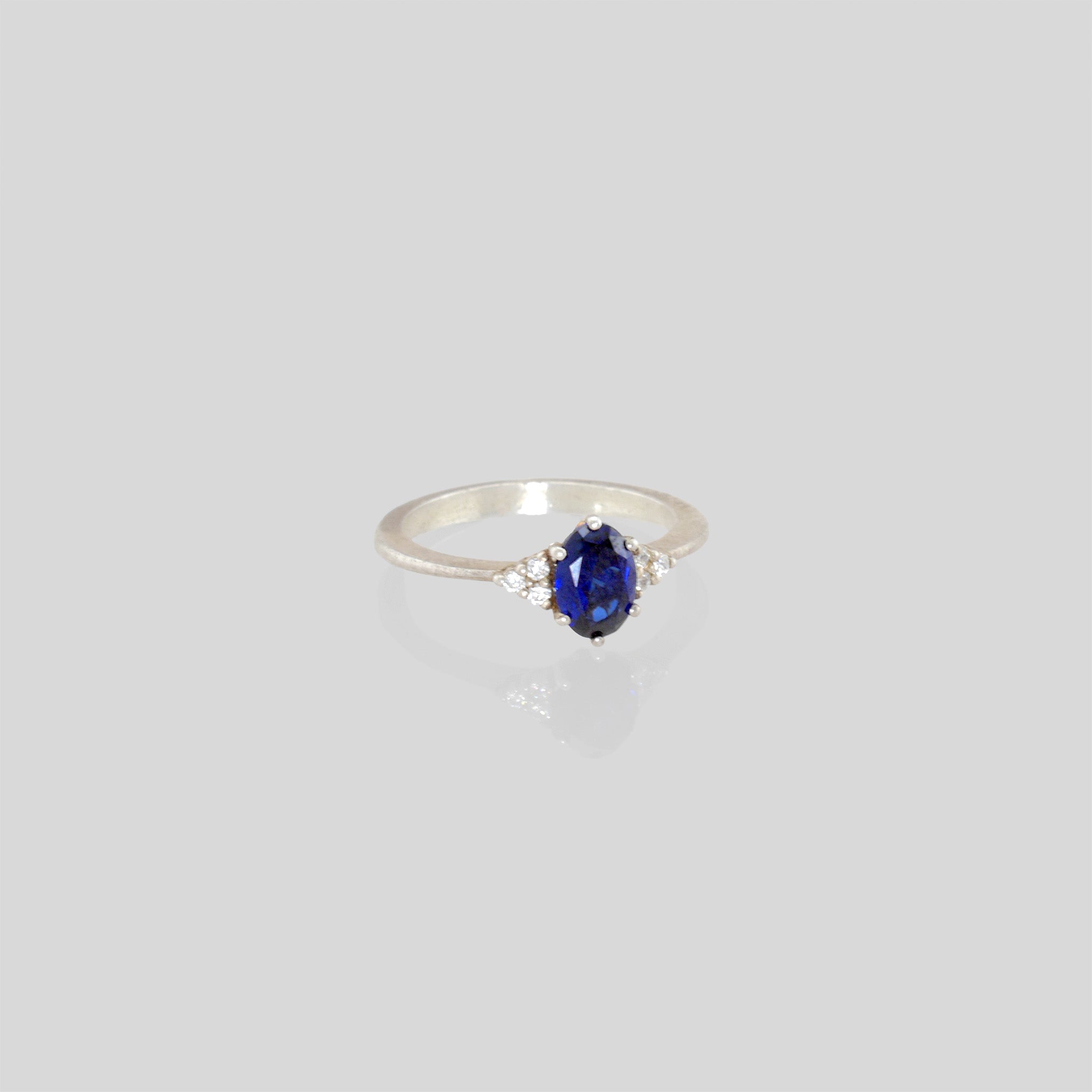 White Gold engagement ring featuring an oval central Sapphire and three dainty Diamonds on each side, offering timeless elegance with added sparkle.