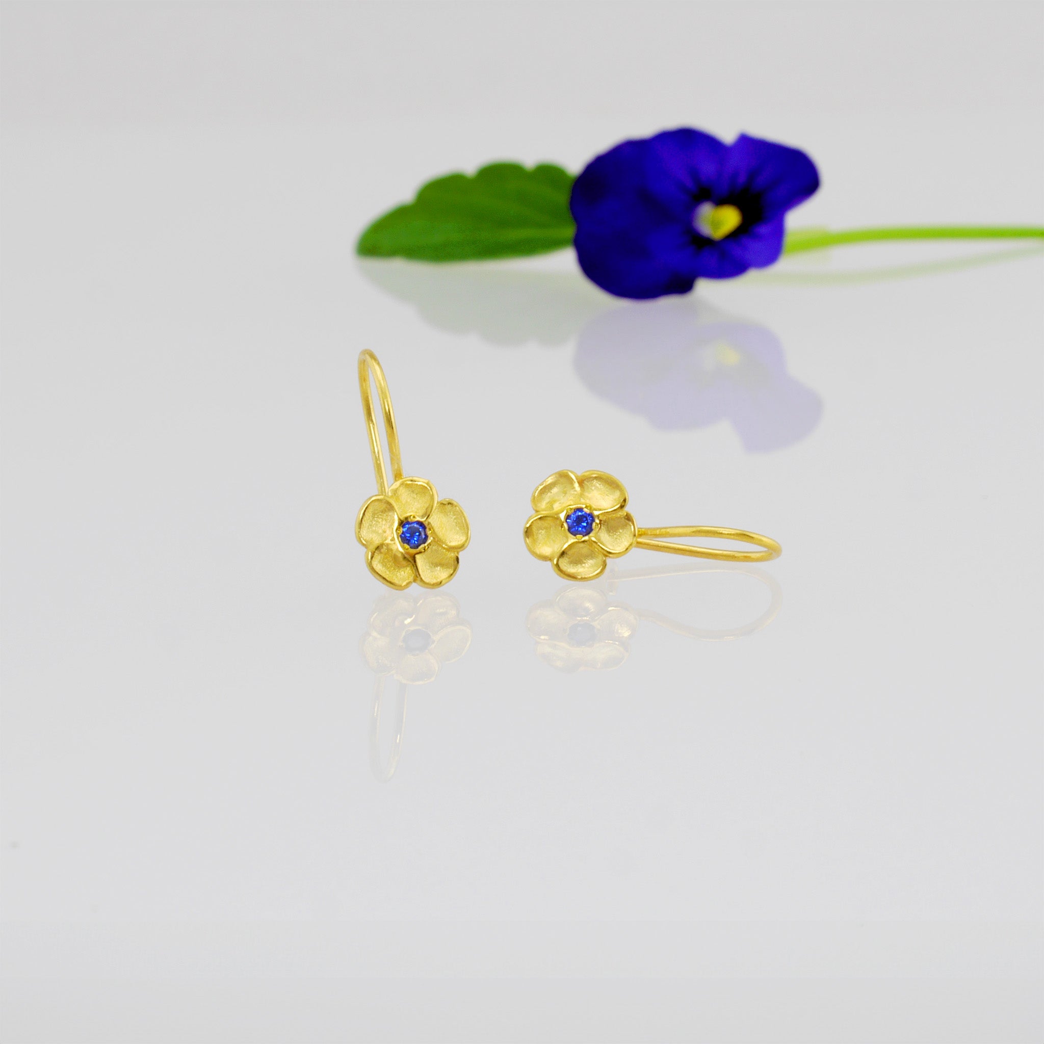 Delicate yellow gold flower drop earrings with a sparkling Sapphire center lying on a bright shining surface with a blue flower in the background. 