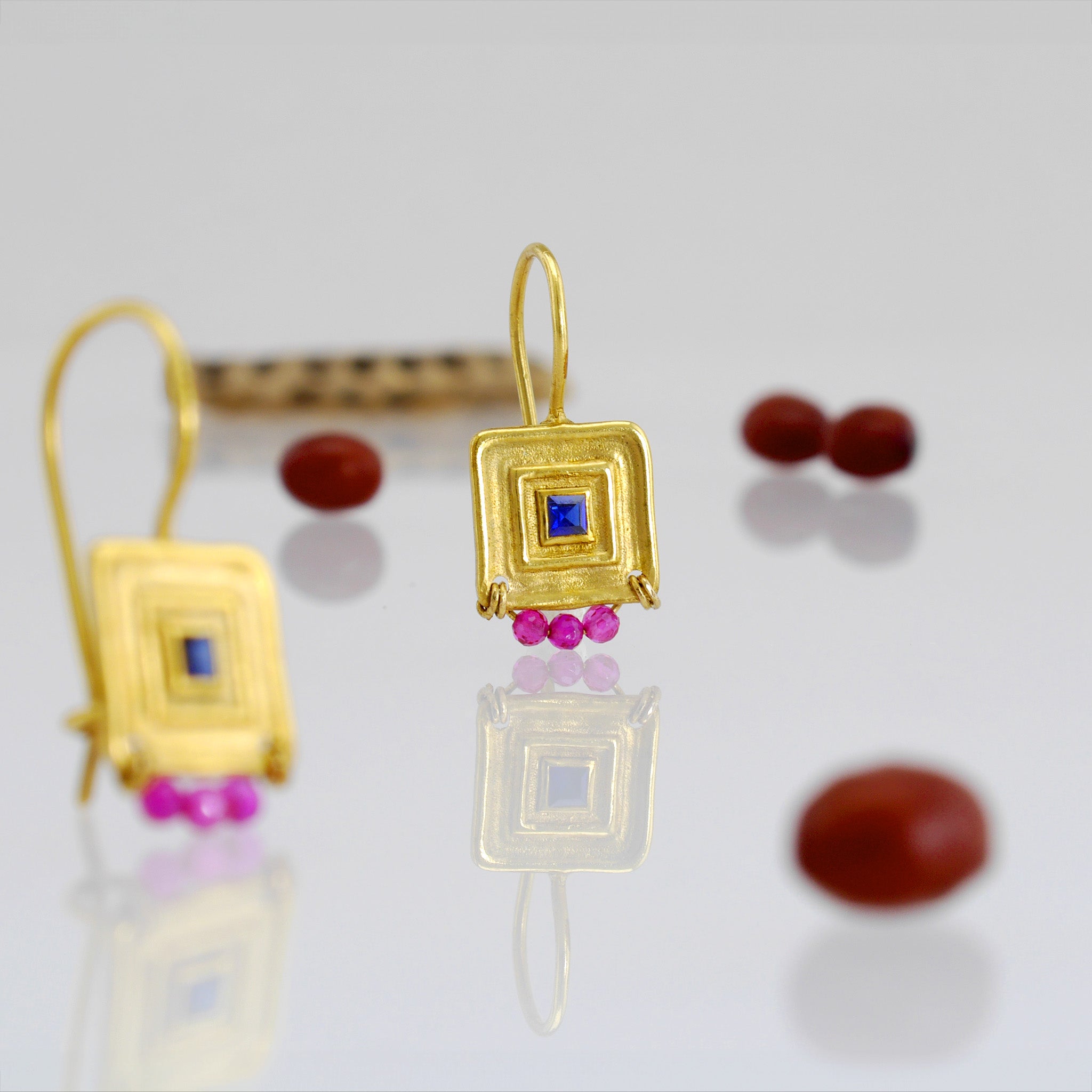 Close up of Unique gold square drop earrings featuring a square sapphire gemstone framed by a line pattern, with three hand-woven ruby beads at the bottom for an elegant Egyptian vibe.