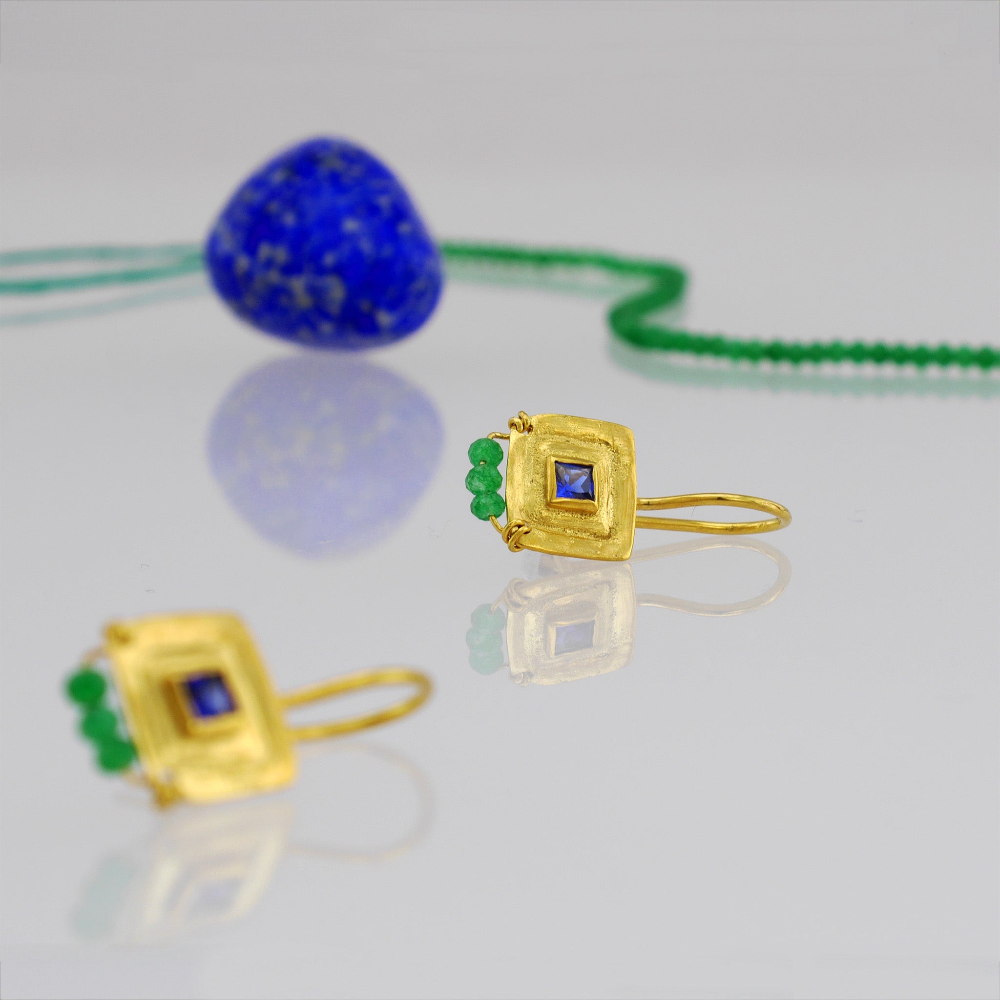 close up of Pharaohs - Gold square drop earrings with square sapphire gemstone, handmade square pattern, and three hand-woven Emerald beads and emerald beads necklace in the background