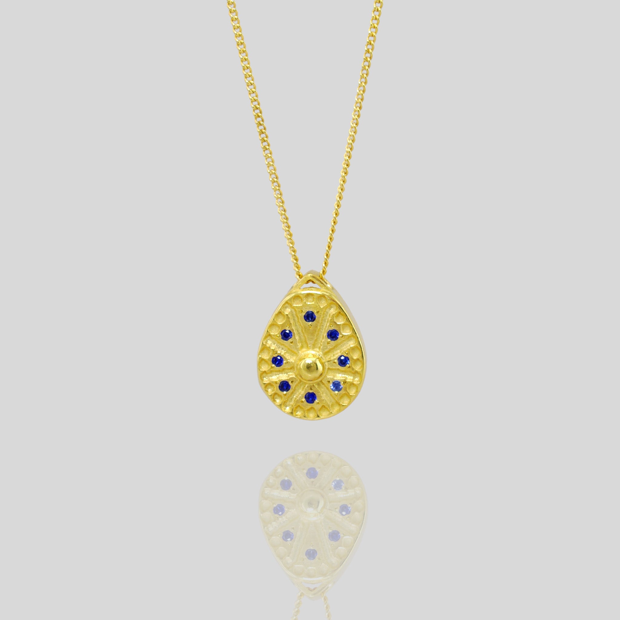 Close up of 18k Gold pendant resembling a Sundial clock, accented with small Sapphires between the edges of each clock hand. A luxurious and timeless addition to any collection.