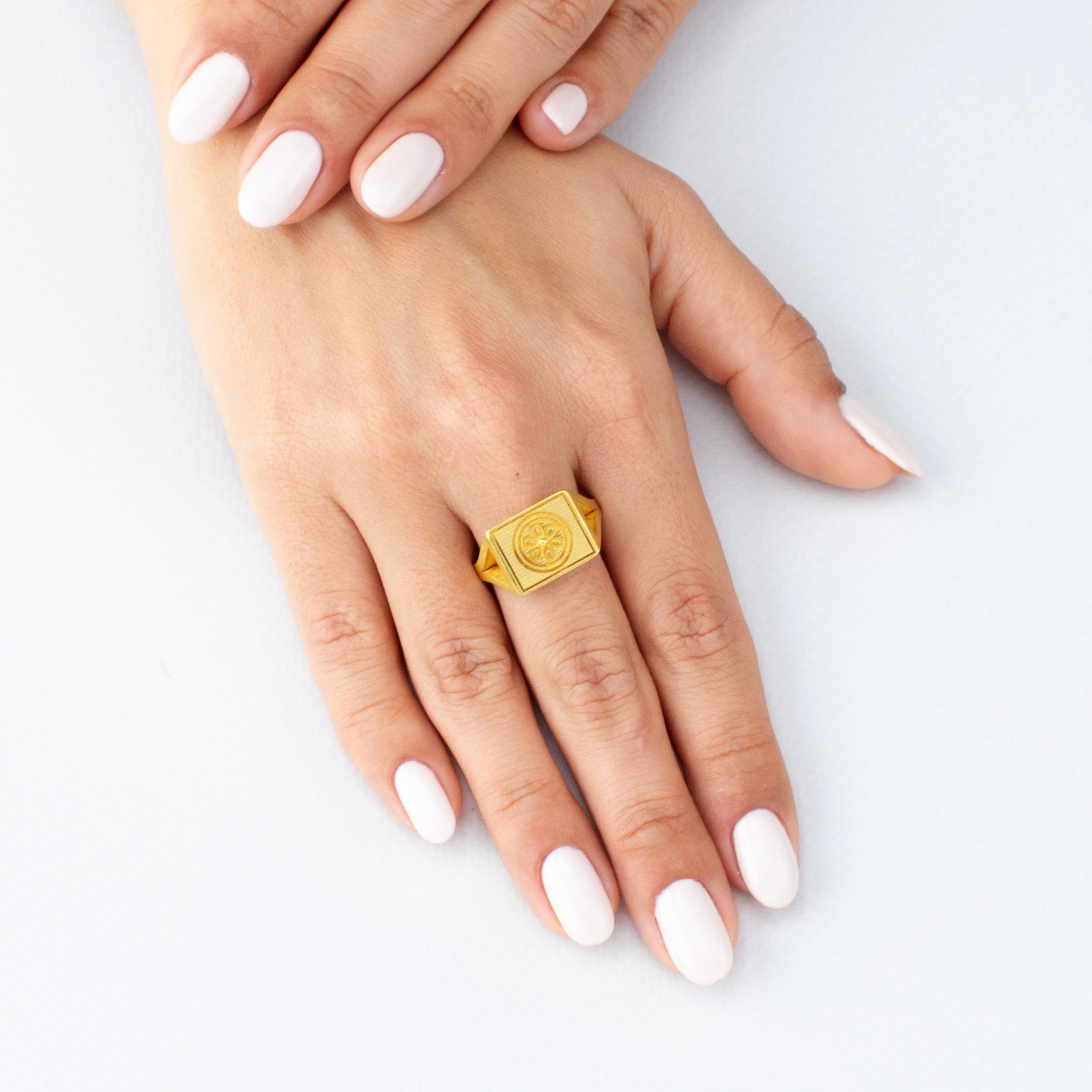 Model displaying an Elegant 18k gold signet ring with a square top featuring an embossed flower design, showcased against a reflective white background.