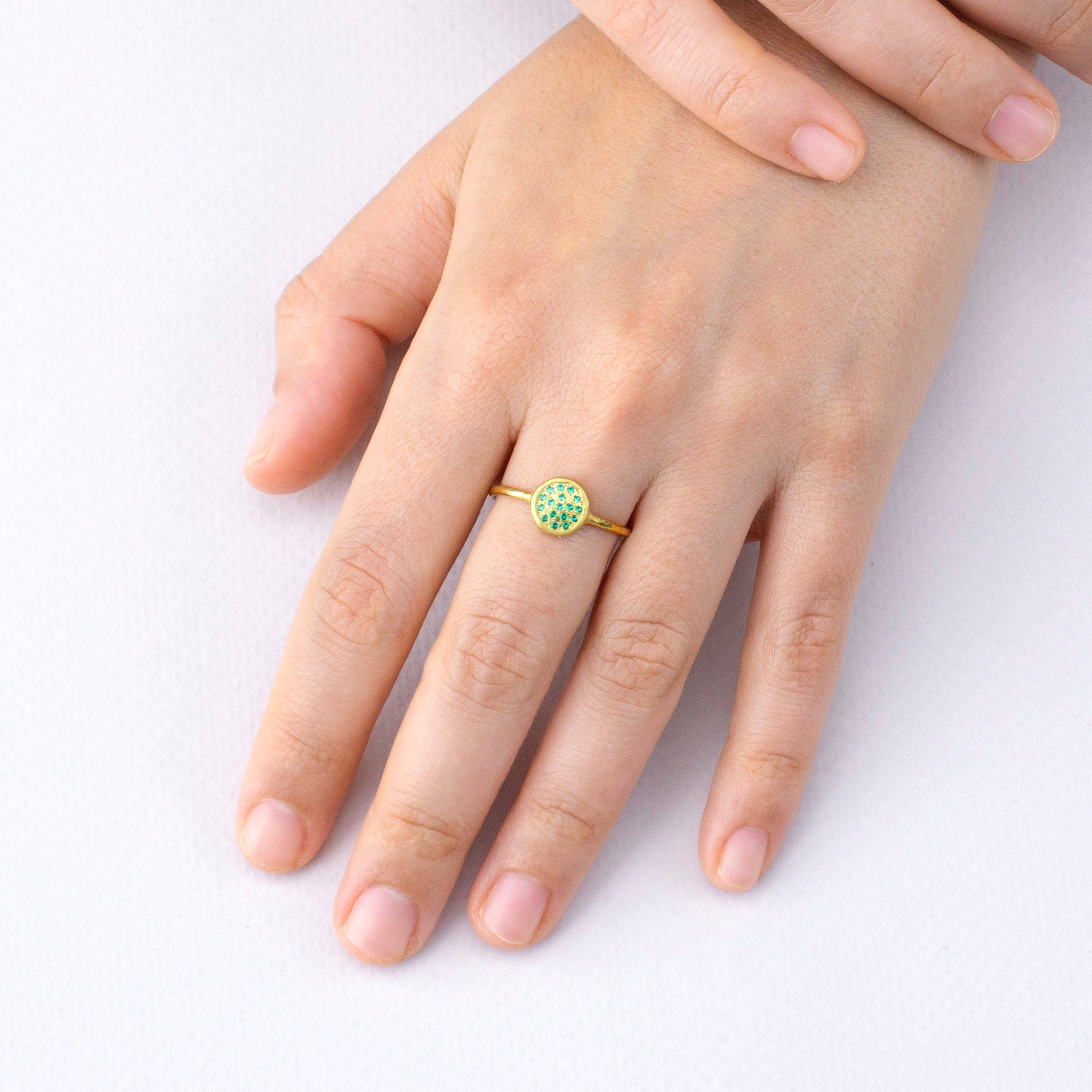 Model's hand elegantly displaying the Starlit gold ring, its circular emerald-encrusted plate shimmering brilliantly, reminiscent of a clear, starry night.